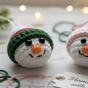May include: Two crocheted snowman ornaments. One snowman wears a green and red hat, the other a pink hat. Both have black eyes, orange carrot noses, and stitched smiles. White yarn and a tag that says "Handmade with Love" are also visible.