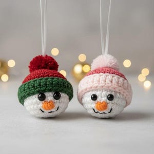 May include: Two handmade snowman ornaments. One snowman has a red and green hat with a red pom-pom, the other has a pink and white hat with a pink pom-pom. Both have black eyes and orange carrot noses.
