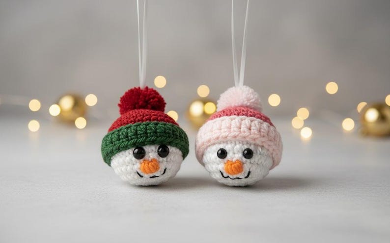 May include: Two handmade snowman ornaments. One snowman has a red and green hat with a red pom-pom, the other has a pink and white hat with a pink pom-pom. Both have black eyes and orange carrot noses.