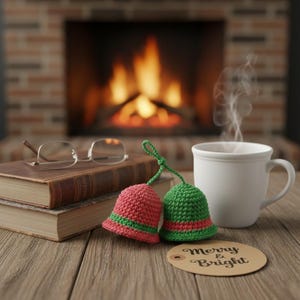 May include: Two crocheted bell ornaments, one red and one green, with green and red stripes, sit on a wooden table next to a white mug and a tag that says "Merry & Bright". A pair of glasses rests on a stack of books, with a fireplace in the background.