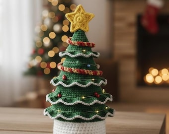 May include: A handcrafted crochet Christmas tree with a yellow star topper. The green tree has white trim, a red and gold garland, and small lights. It sits on a white base, perfect for holiday decor.