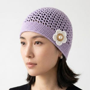 May include: A lavender crochet hat with a floral embellishment. The hat features an open weave design and a solid band at the base. A white flower with a gold beaded center is attached to the side.