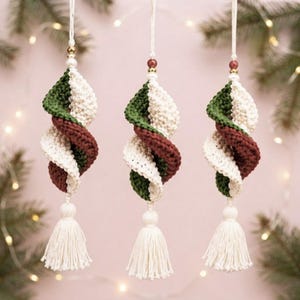 Op de afbeelding: Drie hangende ornamenten met een spiraalvormig haakontwerp in afwisselend crème, bosgroen en donker bordeauxrood garen. Elk ornament is bekroond met een kleine kraal en afgewerkt aan de onderkant met een crèmekleurig garenkwastje.