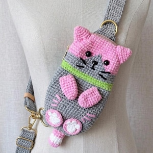 May include: A handmade crochet crossbody bag shaped like a cat. The bag features a pink head, gray body, and pink paws. It has black whiskers and eyes, with a green stripe. The bag has a gray strap with gold-colored hardware.