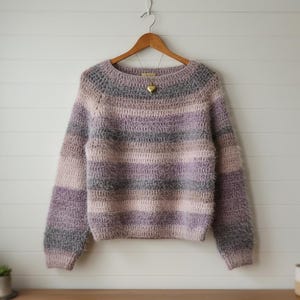 Op de afbeelding: Een handgemaakte, gestreepte trui in tinten lavendel, grijs en roze hangt aan een houten hanger. De trui heeft een ronde halslijn en lange mouwen. Een gouden hartvormige hanger hangt aan de halslijn.