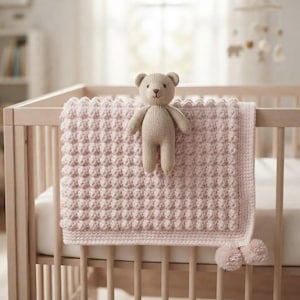 Puede incluir: Una manta de bebé tejida en color rosa claro con un patrón de puntos texturizados y un borde sólido, que presenta dos pequeños pompones en las esquinas inferiores. Un pequeño oso de peluche tejido en color marrón claro está en el centro.