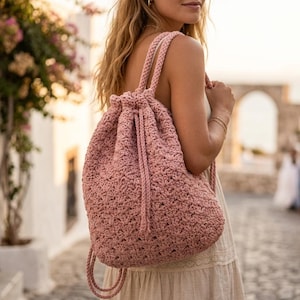 Puede incluir: Una mochila de ganchillo rosa polvoriento con cierre de cordón y correas trenzadas para los hombros. La bolsa tiene un patrón de puntada texturizada y una forma redondeada. Las correas son ajustables. La bolsa está hecha de un material suave, posiblemente de algodón.
