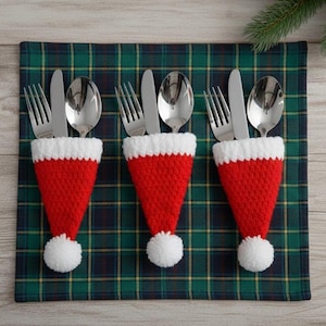 Puede incluir: Tres portacubiertos con forma de gorro de Papá Noel, hechos de tejido de punto rojo y blanco, cada uno con un tenedor, un cuchillo y una cuchara. Los portacubiertos están sobre un mantel individual a cuadros verdes y azules, con una ramita de hoja perenne en el fondo.