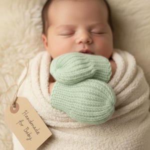 May include: A newborn baby wrapped in a soft, cream-colored blanket, wearing mint green knitted mittens. A brown tag with the words "Handmade for Baby" hangs from the blanket. The baby is sleeping peacefully.