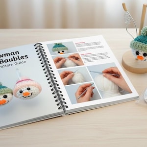 May include: An open spiral-bound book titled "Snowman Head Baubles Crochet Pattern Guide" with images of crochet snowmen. A finished snowman bauble with a green and blue hat sits on a wooden stand. A small bag of white yarn and crochet tools are also present.