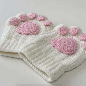 Może przedstawiać: Para białych, szydełkowych rękawiczek bez palców z różowymi detalami w kształcie poduszek i palców u łap. Rękawiczki wykonane są z miękkiej, teksturowanej przędzy i mają przypominać kocie łapy. Rękawiczki są prawdopodobnie przeznaczone do celów nowatorskich lub kostiumowych.