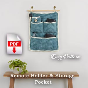 Op de afbeelding: Een turquoise gehaakte afstandsbediening en opbergvak met meerdere vakken, hangend aan een houten deuvel. De houder heeft een crèmekleurige rand en staat op een witte muur. De tekst "Easy Pattern" is zichtbaar.