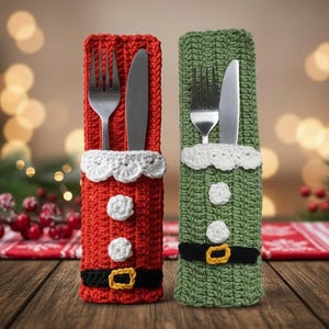 Op de afbeelding: Twee gehaakte kerstbestekhouders. Eén is rood met witte rand, knopen en een zwarte riem met een gouden gesp. De andere is groen met witte rand, knopen en een zwarte riem met een gouden gesp. Elk bevat een vork en een mes.