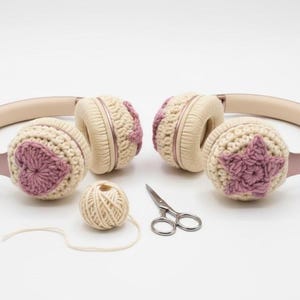Könnte beinhalten: Cremefarbene Kopfhörer mit gehäkelten Ohrmuscheln mit rosa Blumenmustern. Ein Wollknäuel und eine kleine Schere sind ebenfalls vorhanden. Die Kopfhörer haben ein hellrosa Kopfband und Ohrpolster.