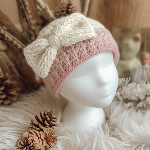 Op de afbeelding: Een handgemaakte gehaakte muts in zachtroze en crèmekleur, met een grote strik detail. De hoed wordt getoond op een witte mannequin hoofd, met de ingewikkelde steek en het ontwerp. Dit gezellige winteraccessoire is perfect om een vleugje handgemaakte charme toe te voegen.