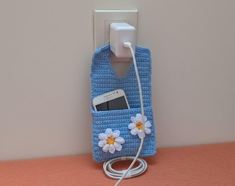 May include: A light blue crochet phone holder with two white daisy appliques. A white smartphone is in the pocket, and a white charging cable is connected to a wall charger. The holder is hanging on a wall.