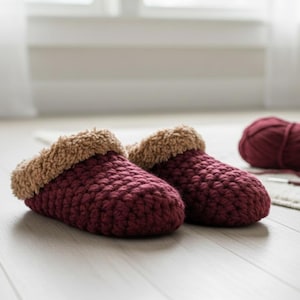 May include: A pair of burgundy crochet slippers with a soft, tan-colored faux fur lining. The slippers are handmade and sit on a light-colored wooden floor. A ball of burgundy yarn is visible in the background, suggesting a cozy, handmade theme.