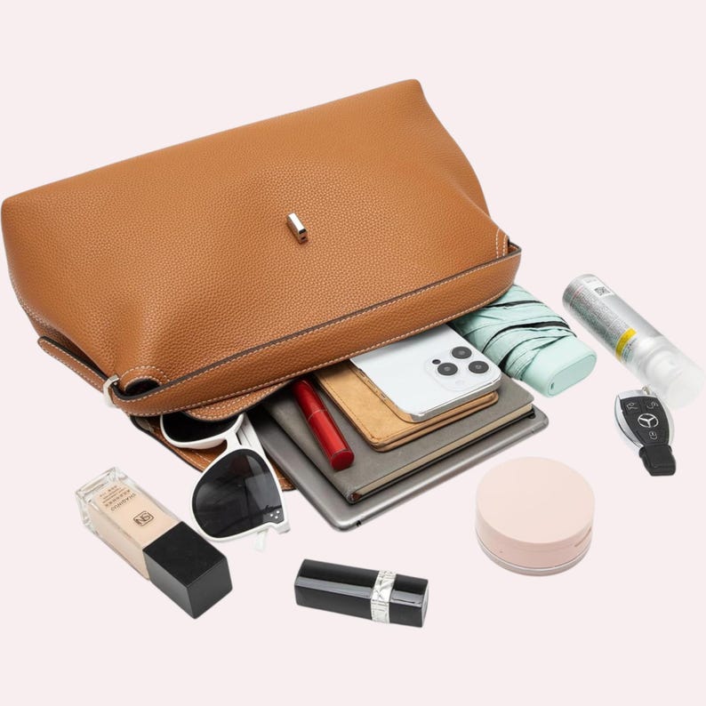 May include: A tan leather handbag with contents spilling out. Items include a smartphone, sunglasses, lipstick, a compact, a small umbrella, and a car key fob. The bag is open, revealing its interior.