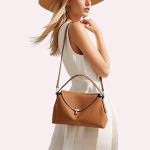 May include: A tan leather handbag with a top handle and a shoulder strap. The bag has a flap closure with a silver-tone metal accent. The bag is carried by a person wearing a white dress and a straw hat.