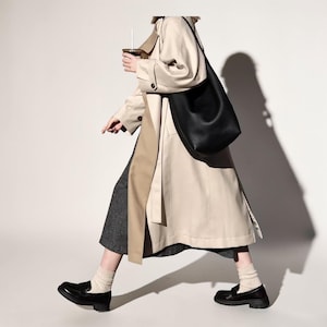 May include: A black leather shoulder bag is carried over the shoulder of a person wearing a beige trench coat and black loafers. The person is holding a coffee cup with a straw. The outfit includes a gray skirt and cream-colored socks.