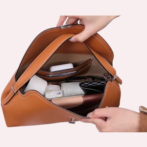 May include: A brown leather handbag with white stitching and a top handle. The bag is open, revealing multiple compartments and contents, including a white case, a jar, and a wallet. The bag has a silver clasp.