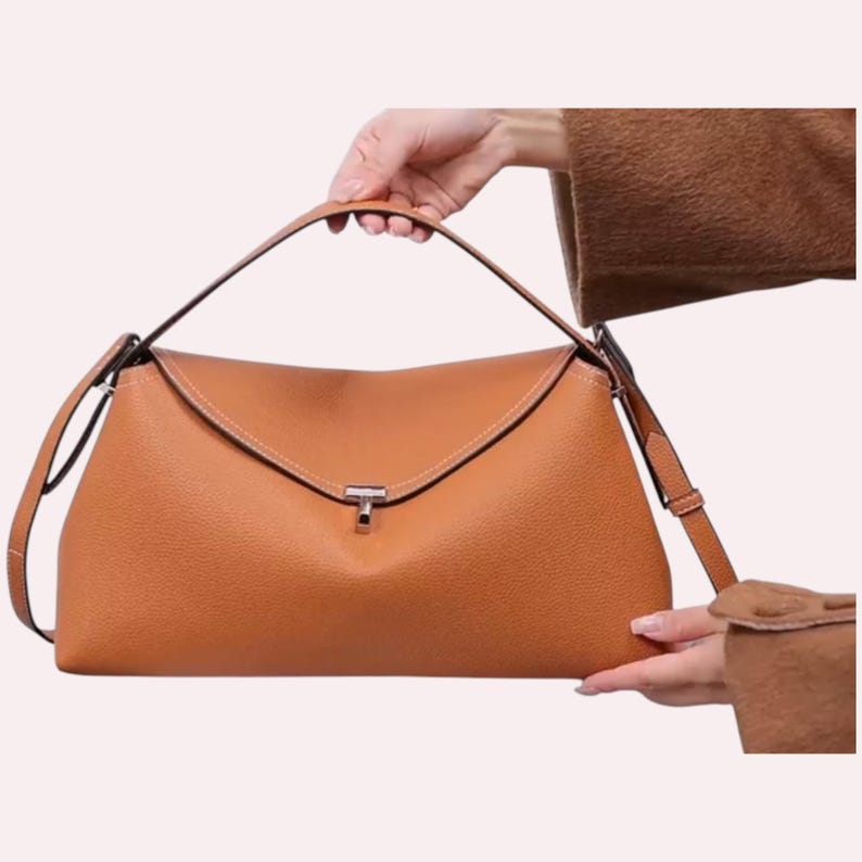 May include: A tan leather handbag with a top handle and a detachable shoulder strap. The bag features a flap closure with a silver-tone clasp and white stitching details. The bag is held by a person.