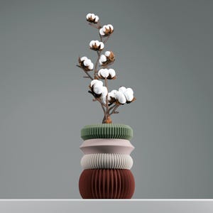Può includere: Un vaso decorativo con un ramo di cotone. Il vaso è composto da quattro sezioni impilate e strutturate in tonalità di verde, rosa, bianco e marrone. Il ramo di cotone presenta batuffoli di cotone bianchi e steli marroni.