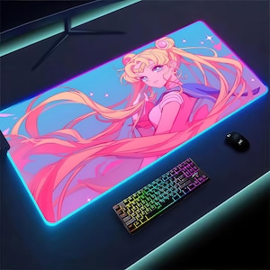 Puede incluir: Alfombrilla de ratón rectangular con una vibrante ilustración de anime de un personaje con largo cabello rubio y un atuendo rosa. La alfombrilla tiene una combinación de colores azul y rosa con un borde azul brillante. También se ven un teclado y un ratón negros.