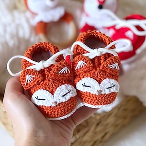 Può includere: Un paio di scarpine da neonato all'uncinetto arancioni a forma di volpe, con dettagli bianchi e lacci bianchi. Le scarpine presentano facce di volpe con musetti bianchi, occhi neri e dettagli delle orecchie bianche. Altri articoli per bambini sono sullo sfondo.