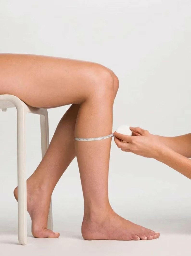 May include: A white retractable measuring tape wrapped around a lower leg to measure circumference. The tape features clear black markings in inches, showing a measurement of approximately 12 inches around the calf area of the lower leg.