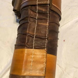 May include: A cylindrical leather roll featuring alternating bands of dark brown and tan leather. The surface displays a series of horizontal ridges and is secured with a vertical strap and buckle closure. White stitching runs along the seams.