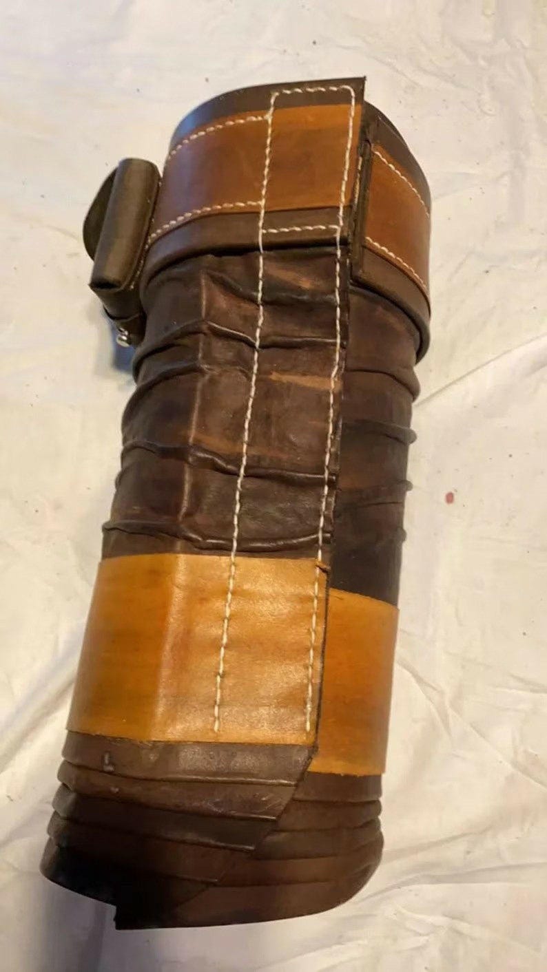 May include: A cylindrical leather roll featuring alternating bands of dark brown and tan leather. The surface displays a series of horizontal ridges and is secured with a vertical strap and buckle closure. White stitching runs along the seams.