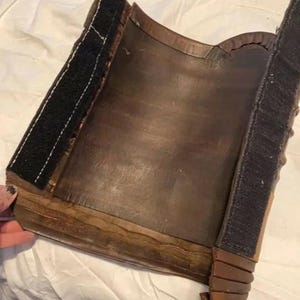 May include: A curved, dark brown leather piece featuring two vertical strips of black hook-and-loop fastener along the edges. The leather has a smooth, worn texture with visible stitching and layered, overlapping segments at the bottom corner.