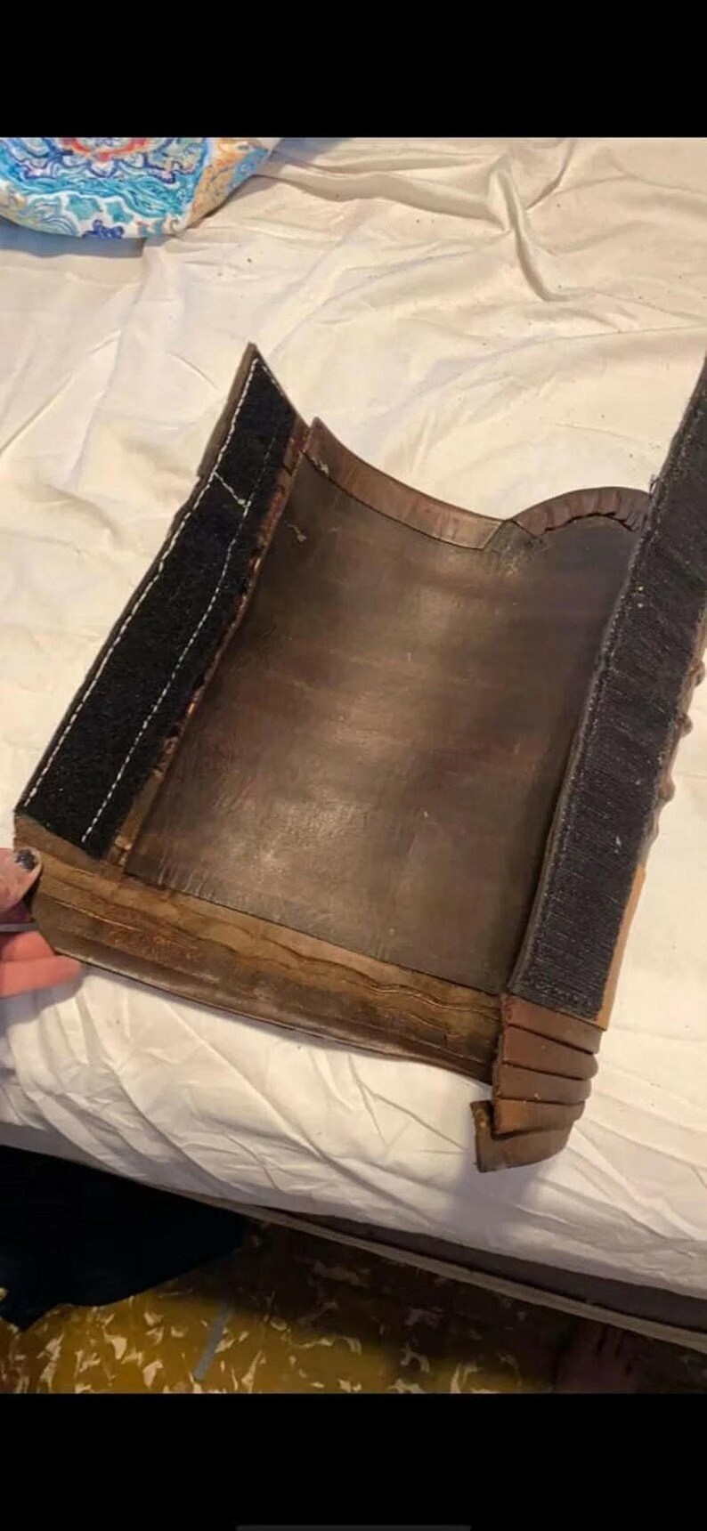 May include: A curved, dark brown leather piece featuring two vertical strips of black hook-and-loop fastener along the edges. The leather has a smooth, worn texture with visible stitching and layered, overlapping segments at the bottom corner.