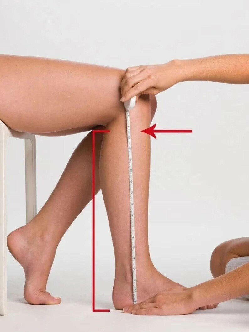 May include: A white measuring tape is positioned vertically along the lower leg, extending from the base of the heel to the bottom of the knee. A red arrow points horizontally toward the knee area, indicating the measurement point for the lower leg length.
