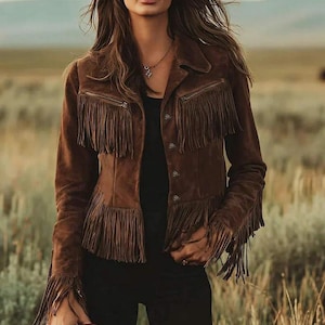 May include: A brown suede jacket with fringing on the chest, sleeves, and hem. The jacket features a button-up front and two zipped pockets. The model is wearing a black top and trousers. The background is a field of tall grass.