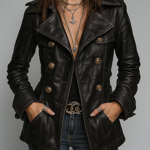 Peut inclure: Veste en cuir noire à double boutonnage avec boutons dorés. La veste a un col cranté, des poches et une ceinture. Le modèle porte un jean bleu et des colliers en argent.