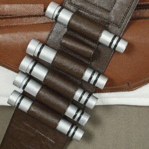 May include: A brown leather bandolier featuring a series of silver cylindrical capsules with black ring details. The bandolier is positioned diagonally across a brown leather strap and a white fabric base, showcasing a structured design.