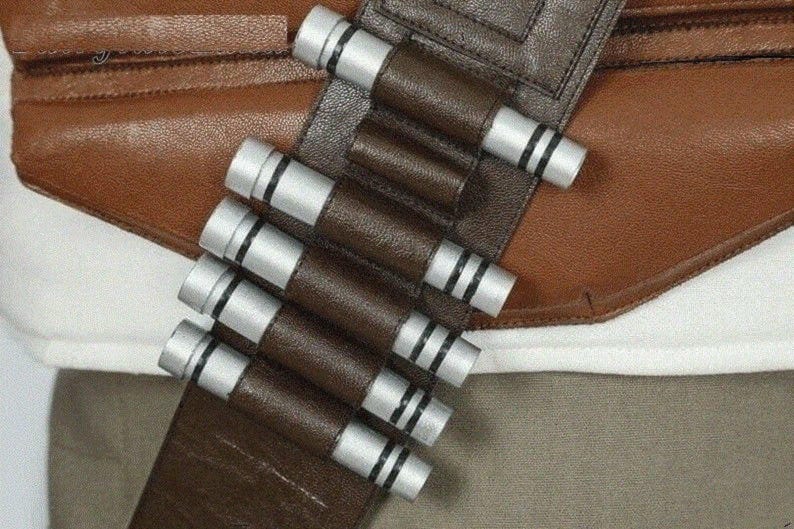 May include: A brown leather bandolier featuring a series of silver cylindrical capsules with black ring details. The bandolier is positioned diagonally across a brown leather strap and a white fabric base, showcasing a structured design.