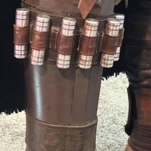 May include: A brown leather leg armor piece featuring a horizontal strap holding five cylindrical silver metal canisters. The armor has a structured, layered design with a dark brown finish and a weathered appearance, secured with a wide leather band.