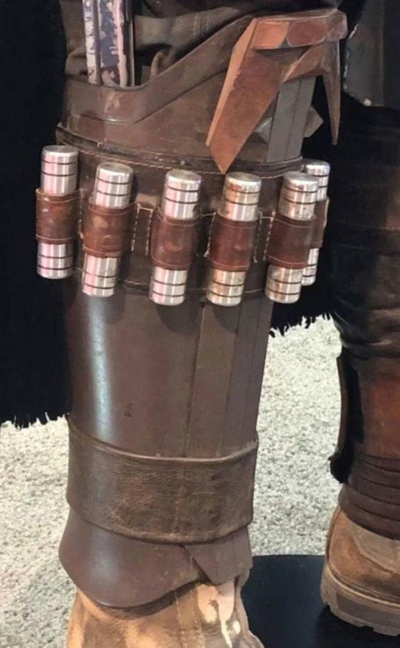 May include: A brown leather leg armor piece featuring a horizontal strap holding five cylindrical silver metal canisters. The armor has a structured, layered design with a dark brown finish and a weathered appearance, secured with a wide leather band.