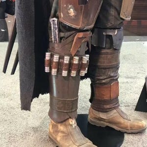 May include: A costume featuring brown leather-like leg armor with metallic accents, including a row of small silver cylinders attached to the shin guard. The outfit includes weathered tan boots, dark trousers, and a long black cape, creating a detailed ensemble.