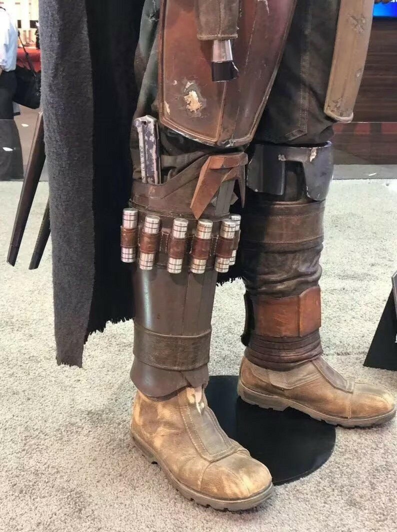May include: A costume featuring brown leather-like leg armor with metallic accents, including a row of small silver cylinders attached to the shin guard. The outfit includes weathered tan boots, dark trousers, and a long black cape, creating a detailed ensemble.