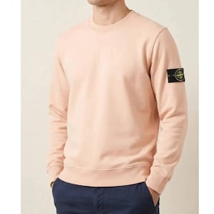 Op de afbeelding: Een licht perzikkleurige sweatshirt met een ronde hals en lange mouwen. Een kleine zwarte vierkante patch met een geel embleem bevindt zich op de linkermouw. De sweatshirt is gemaakt van zacht materiaal en geschikt voor vrijetijdskleding.