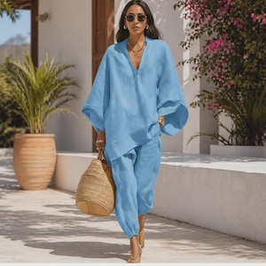 May include: A light blue linen two-piece outfit. The top has a V-neck and wide sleeves. The pants are a cropped, jogger style. A woven tote bag is carried. The person is wearing sunglasses and gold-colored jewelry. The outfit is suitable for warm weather.