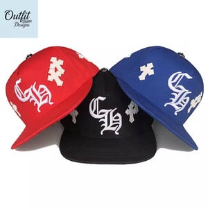 May include: Three baseball caps in red, black, and blue. Each cap features white embroidered lettering and cross designs. The caps have a flat brim and a structured crown. The brand name "Outfit Bloom Designs" is in the top left corner.