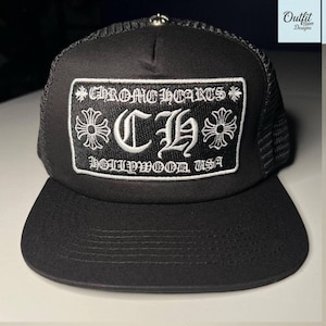 Puede incluir: Gorra de camionero negra con visera curva y parte trasera de malla. El panel frontal presenta un parche bordado blanco con el texto "CHROME HEARTS HOLLYWOOD USA" y elementos florales. Un botón plateado está en la parte superior.
