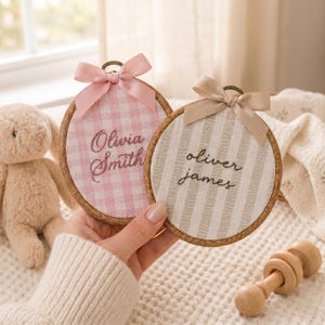 May include: Two personalized embroidered hoops with names, one pink gingham and one striped, each with a ribbon bow. The names "Olivia Smith" and "Oliver James" are stitched in cursive. The hoops are held in a hand.