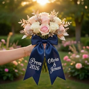 Puede incluir: Un ramo de rosas rosas y blancas con detalles plateados, atado con una cinta azul marino. La cinta tiene el texto "Prom 2026" y "Olivia & Jonas" en escritura blanca. El fondo es un jardín borroso.