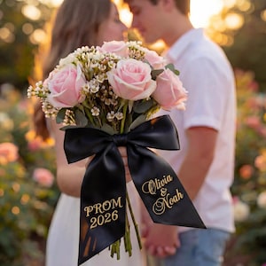 Puede incluir: Un ramo de rosas rosas y flores blancas atado con una cinta de satén negra. La cinta tiene texto dorado que dice "PROM 2026" y "Olivia & Noah". El fondo muestra una pareja borrosa.